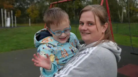 A woman is holding a boy in her arms. They are both smiling at the camera and stand in front of some swings 