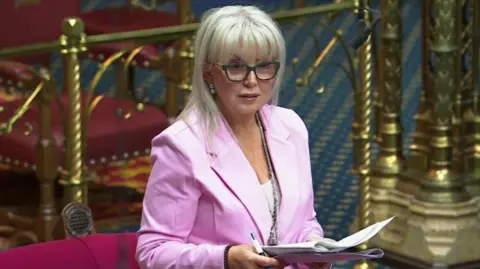 Baroness Helen Newlove, with long blonde hair and dark glasses, wears a pink suit as she addresses the House of Lords. She is holding some papers and a pen.