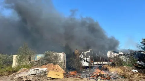 A pile of burning rubbish and sheds surrounded by thick black smoke which is filling the sky