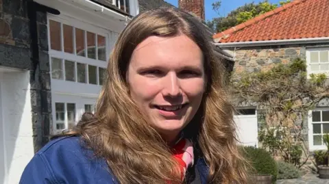 A man with long blonde hair, wearing a blue jacket and pink and red neckscarf smiles at the camera. The background is houses and windows, with foliage growing on the walls.
