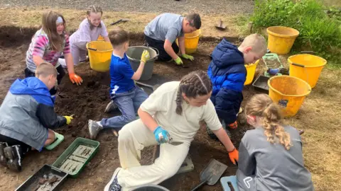 Eight school children kneel on the ground as they dig away at a patch of soil. They have each got trowels and a yellow bucket as they excavate the dirt. 