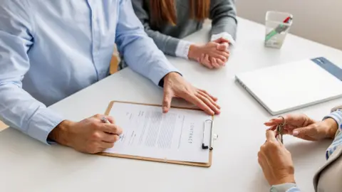 Three people sitting around a white table. None of their faces are shown. One is holding a set of keys, another, wearing a blue shirt, is signing a contract. A third person at the top of the image is wearing a grey top over a white shirt and has her hands clasped. 