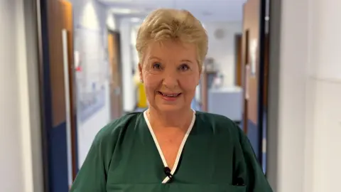 A women in her 70s with short blonde hair in green nurses scrubs. She is stood in front some doors to a ward, a sign above them says 'Welcome to Carey'