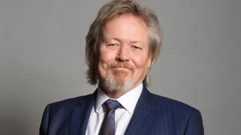 UK Parliament Giles Watling wearing a dark blue, pin-striped suit, white shirt and dark blue patterned tie. He is standing against a plain, grey background. He has a neat goatee beard and light coloured hair that falls over his ears and just reaches his collar.