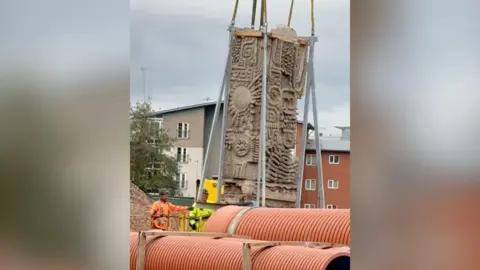The Aztec-style mural being lifted up with two men in bright high-vis jackets standing underneath. Below the mural being lifted is two large orange pipes. In the background are flats.