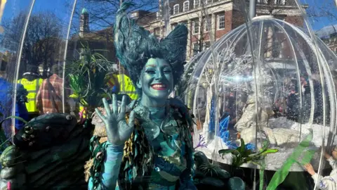 A person in a blue costume, makeup and wig waves at the camera. The snow dome is in the background.