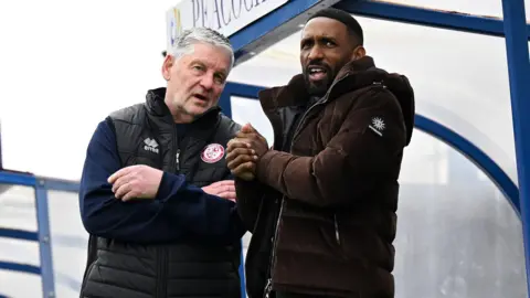 Getty Images Jermaine Defoe in a brown coat standing in a football dugout talking to a man in a Woking FC gillet.