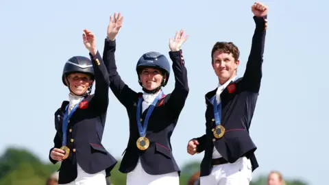 David Davies/PA Great Britain's Rosalind Canter, Laura Collett and Tom McEwen with their gold medals. The trio all have their arms up. Tom is holding his fist up to celebrate. They are all wearing equestrian gear and gold medals. 