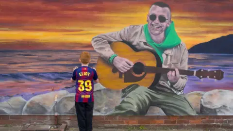 A mural of a man sat on rocks, with the sunset behind, playing a guitar. In front of the wall is a boy is a blue and purple Barcelona shirt with Eliot and 39 on the back in yellow