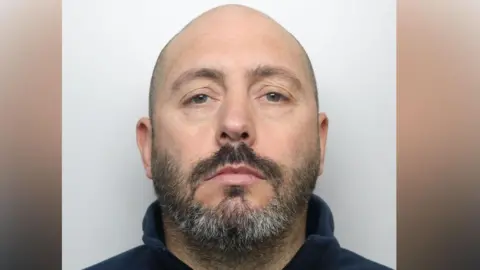 A bald man in his forties, with a greying beard. He is wearing a blue hoodie.