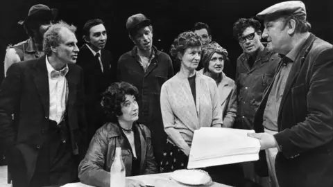 Arthur is stood on the right hand side with a group of male and female actors looking at him while he gives direction in a black and white photo. He is holding a script and is wearing a cap and casual clothes.