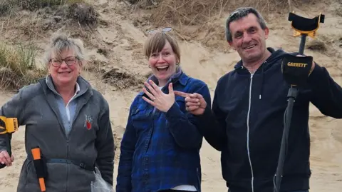 From left to right, Lorna Rawlings who has grey hair and a metal detector,Sarah Stone who brown hair, a delighted expression on her face and is holding up her hand to show a wedding ring, Andy Granr, who has grey hair and is holding a metal detector. 