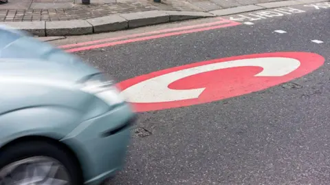 Richmond Council A red circle with a large white letter C is painted on the surface of a road in front of a crossing. A silver-blue car is driving towards the painted C from the left, and is blurred from motion. The pavement with double red lines along the kerb is visible in the background.