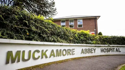 PA Media A sign says Muckamore Abbey Hospital on a low white wall in front of bushes and a red-brick building