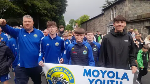 Shows young people wearing blue football tops and carrying a banner reading Moyola Youth