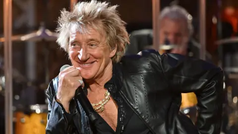 Rod Stewart in a leather jacket and shirt unbuttoned halfway down his chest, holding a microphone and leaning to one side