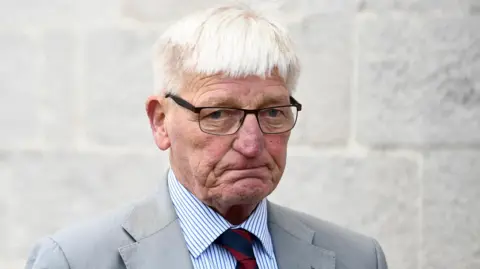 Pacemaker Former Soldier Dennis Hutchings Armagh Court. He has white hair, wearing glasses, a grey blazer, blue and white pin stripe shirt and red and navy tie.