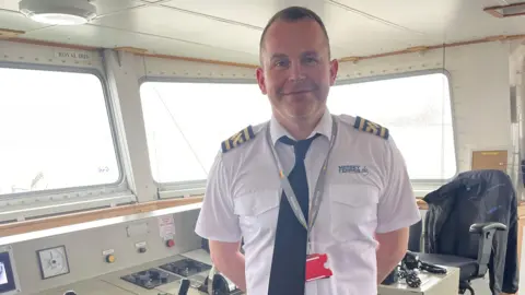 Captain Gary Simpson smiles in the control deck. He wears the Mersey Ferries liveried uniform shirt and tie.