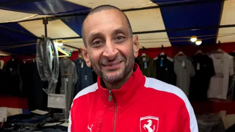 Bob Sidhu sonriendo. Lleva una chaqueta roja. Está en un mercado y hay ropa detrás de él.