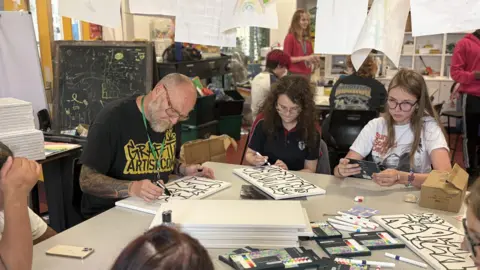 Men and women are sitting on a table holding pens and drawing fonts on canvases. They are sitting in an art room with work drying from the ceilings.