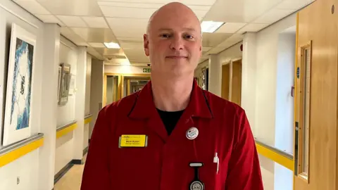 Mark Butler, a bald man, wearing a red matron uniform in a hospital corridor.