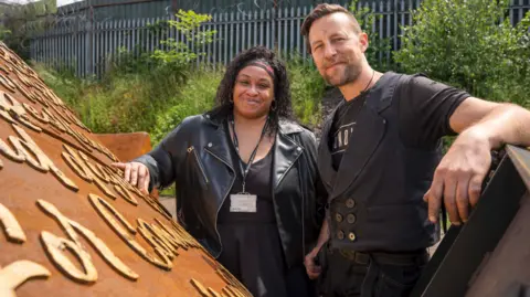 Victoria Murrain (left) and sculptor Luke Perry stand with the Windrush monument which is partially hidden from view in the park. What can be seen is a Corten steel colour with writing on it. The artists stand together behind it both wearing black and smiling at the camera. Victoria has long black hair and a scarf around her head. Luke has short brown hair and a beard. 
