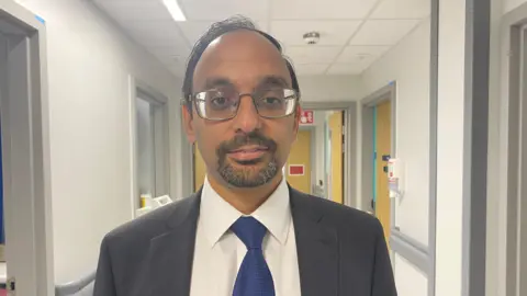 Saurav looking into the camera and smiling slightly. He's wearing a dark grey suit, with a white shirt and a dark blue tie. He's stood in a hospital corridor which has white walls and grey doorframes.