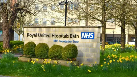 RUH Bath A cement NHS sign outside the RUH in Bath. It is surrounded by bright green grass, blooming daffodils, manicured bushes and several trees. In the background you can see the hospital, a large white building with lots of windows.