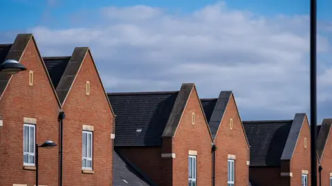 Getty Images The image shows a new build house development estate; the bricks are red and brown and all the houses have one window facing outward. The shy is cloudy and slightly grey.