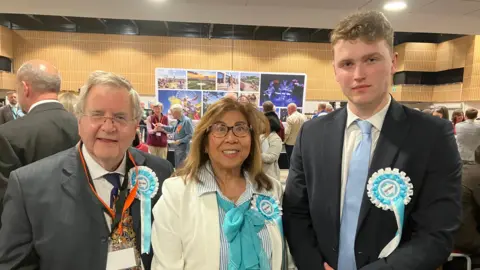 Three people wearing “Reform UK” rosettes stand together in a busy indoor hall, dressed in formal attire. Behind them, groups of attendees gather and converse near a stage area with posters, under bright overhead lighting and a high, wood-paneled interior.