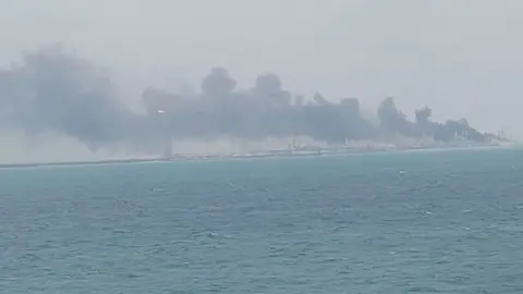 Sailor provided to BBC News Plumes of smoke drift from the right of the photo over buildings along a shoreline. Dark blue sea is in the foreground, and the sky is light blue.