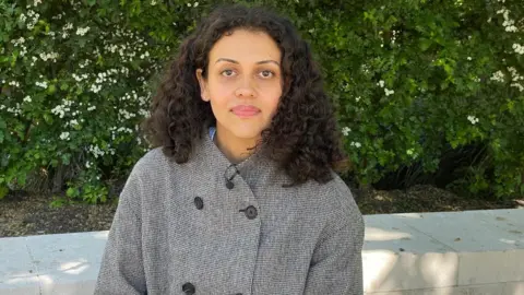 Founder Faustine Petron wearing a grey coat with large black buttons. She has shoulder-length curly black hair and is sitting in the shade beside a low wall and a hedge with white flowers.