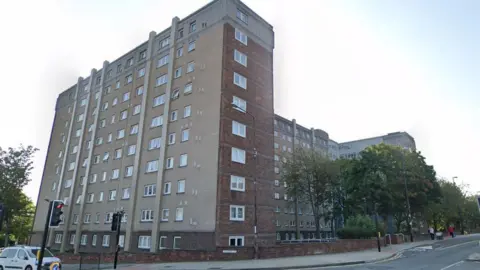 Google Streetview of a 10-storey block of flats. It is a combination of red brick and concrete. On the narrower side there are single windows on each floor. On the longer side there are 10 windows per floor. The block sits on a corner next to a set of traffic lights. 