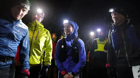 David Miller Photography A picture of several runners standing in the rain in the dark, with head torches on. The main focus of the picture is a woman with a purple waterproof on and a head torch. Her hood is up and she is laughing