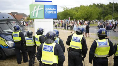 Riot police, pictured from behind, line up alongside a sign indicating a Holiday Inn Express. Disparate groups of people mill about in front of them. 