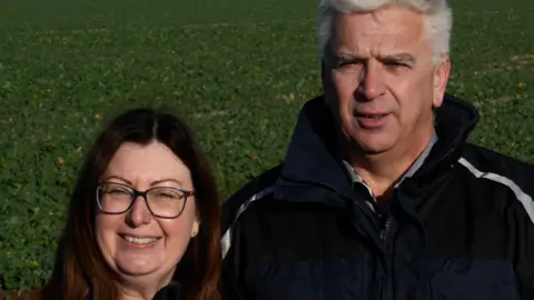 John Fairhall/BBC Susanne Eves, with brown hair and spectacles, standing next to Mark Eves, with grey hair, wearing a navy jacket.