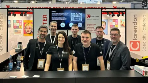 Clarendon Specialty Fasteners Staff all smiling at the camera in a group with black t-shirts on. They are in front of lots of branded banners in a big expo-type event in a warehouse-style building