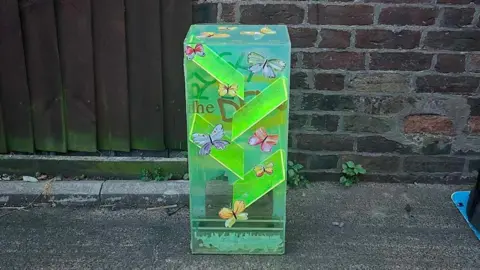 RUSH 2The DEN A luminous plastic money box, with butterfly designs on the front, in front of a brick wall. 