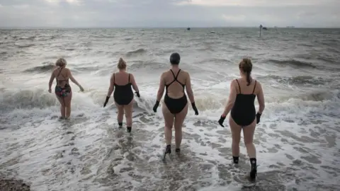 PA Four swimmers in black wetsuits and gloves walk into the sea as the waves crash in. The sky is grey.