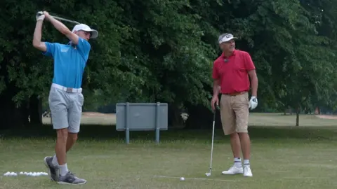 Jamie Niblock/BBC Liam, who is a left-handed golfer, hits a shot with an iron on the range at Dereham Golf Club. His club-mate Andrew smiles as he admires the shot. The pair are hitting shots off the grass with bushes and trees behind.