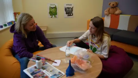 Shaun Whitmore/BBC Woman wearing purple hoodie seated by a craft table next to a teenage girl wearing a white patterned t-shirt. They are making memory jars using coloured sand.