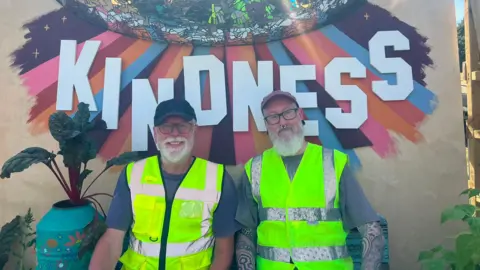 Both men are wearing caps, glasses and hi-vis jackets. They have white beards and are smiling. Behind them in large letters is the word 'kindness' with rainbow colours and a disco ball.
