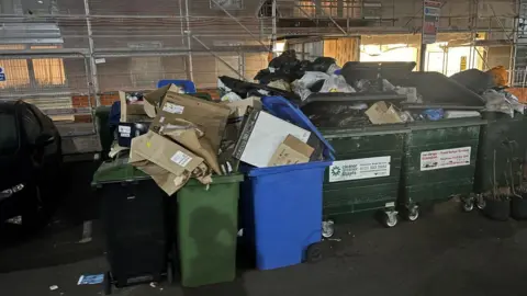 Chris Whittock A row of bins full of waste is lined up alongside recyling bins full of cardboard next to a building with scaffolding.