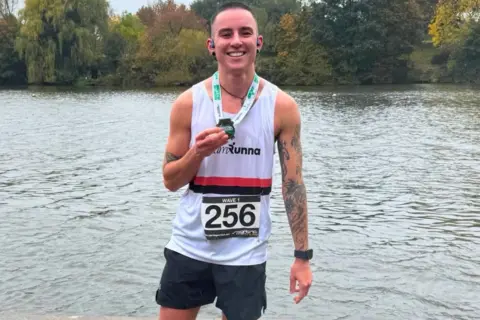 Katie Reynolds is a man standing in front of a lake. He is wearing ongoing clothes and a bib "At 256" And smiling on the camera. He is holding a medal