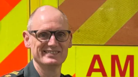 East of England Ambulance Service Simon Chase, a paramedic, smiles at the camera wearing a green paramedic uniform. He is standing in front of an ambulance. 