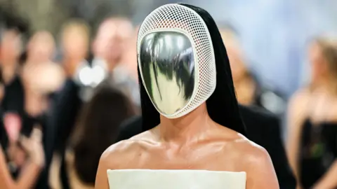 Katy Perry is seen from the shoulders up. She has on a metal and white mask. She has on a white top with bare shoulders and she has long black hair