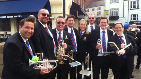 NASUWT Six men, including Tony Thompson, and two women, wearing black blazers and pants and red and blue striped tie, hold brass instruments and recording books as they stand in a town shopping square.
