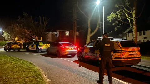 A road with three police cars on it. It is dark and houses are in the background. A PSNI officer is standing with his back to the camera.