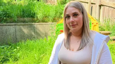 James Grant/BBC A woman in a white top and white hoodie stands in an overgrown back garden. 