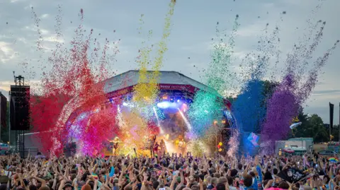 The main stage at Bristol Pride, in front of a huge crowd. Rainbow-coloured confetti can be seen flying from the stage. 